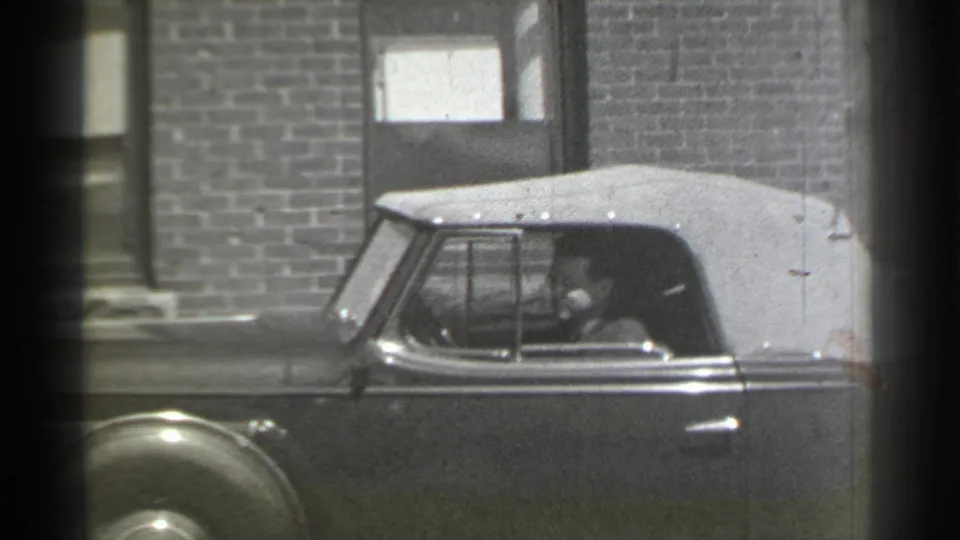 A late-1930s convertible with a light soft top is shown from the side, with the driver seated inside near a brick building.