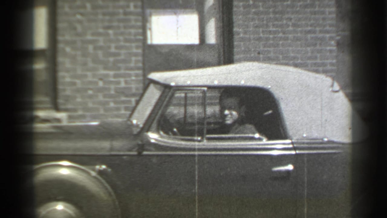 1939 New York home-movie frame showing a soft-top convertible at curbside, brick facade behind it, and a person visible inside the car.