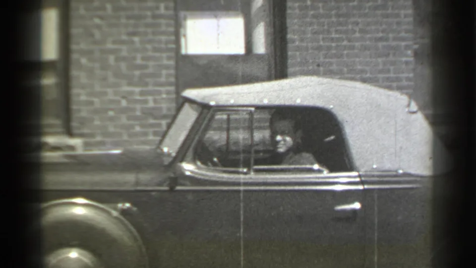 1939 New York home-movie frame showing a soft-top convertible at curbside, brick facade behind it, and a person visible inside the car.
