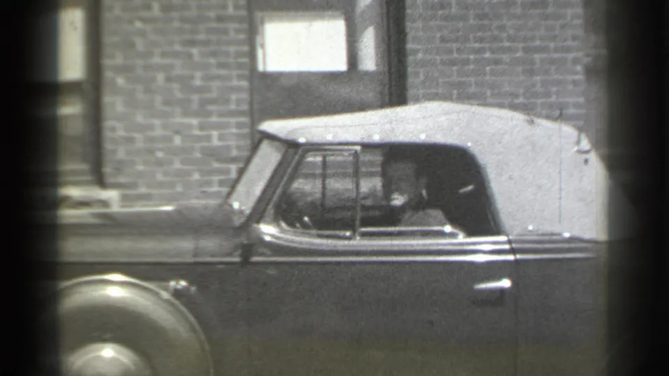 The vintage convertible remains parked in front of the brick exterior, with the seated occupant visible through the side window.