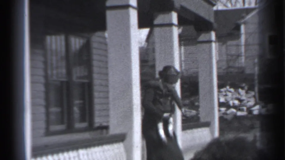 The woman holding the small child moves along the building frontage, with columns, windows, and a raised planting area visible behind her.