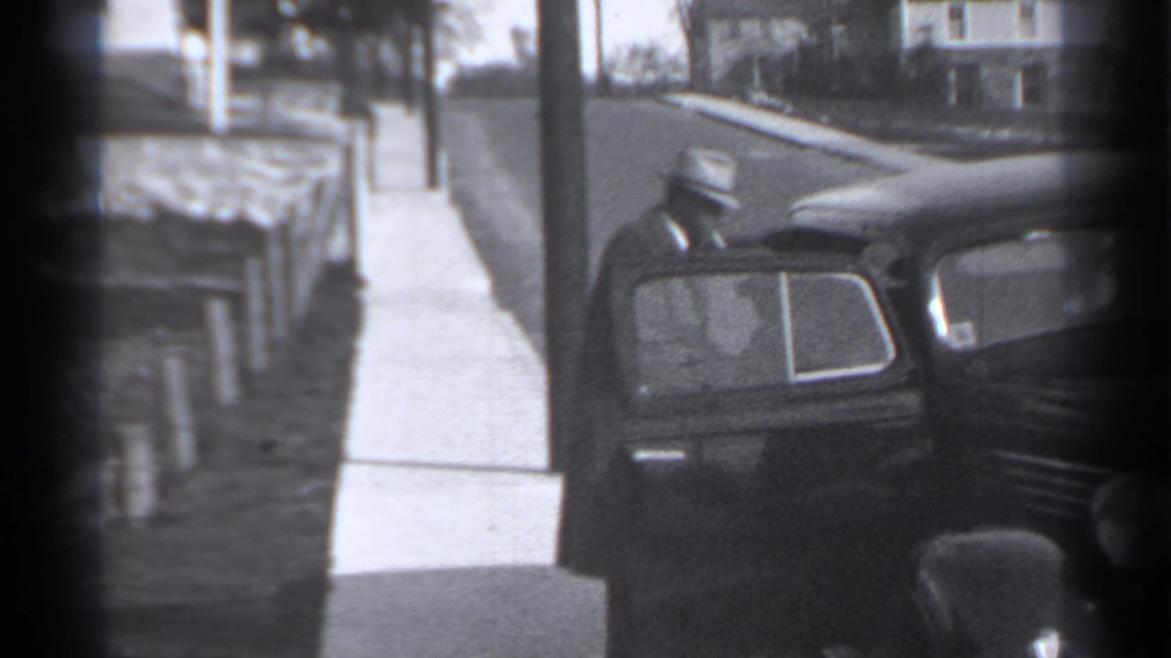 1939 New York home-movie frame showing a dark vintage car by a sidewalk, low fence, utility pole, and houses.