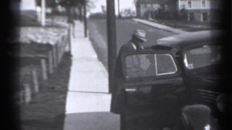 1939 New York home-movie frame showing a dark vintage car by a sidewalk, low fence, utility pole, and houses.