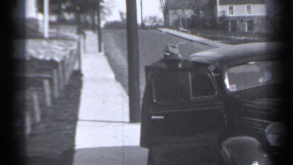 Dark vintage automobile parked on a New York residential street in a soft 8mm frame, with sidewalk and houses behind it.