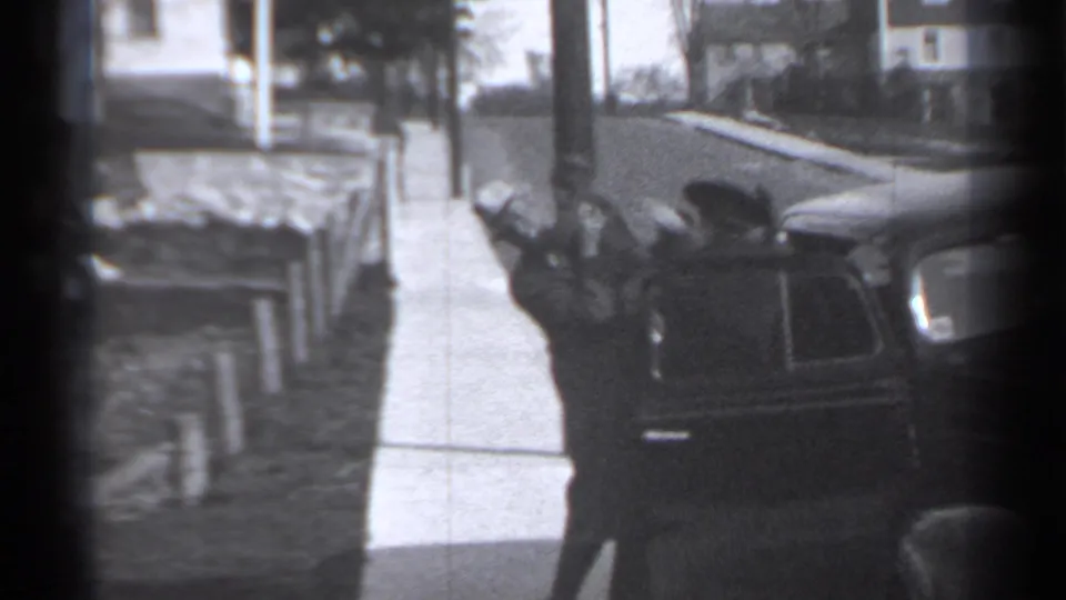 An adult in a suit and hat appears beside the dark car, helping a smaller figure at the curb on a residential New York street.
