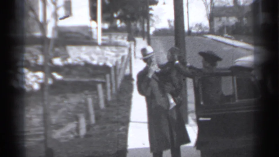 Adult in a long coat and hat stands with a child-sized figure beside a dark vintage car on a residential New York sidewalk.