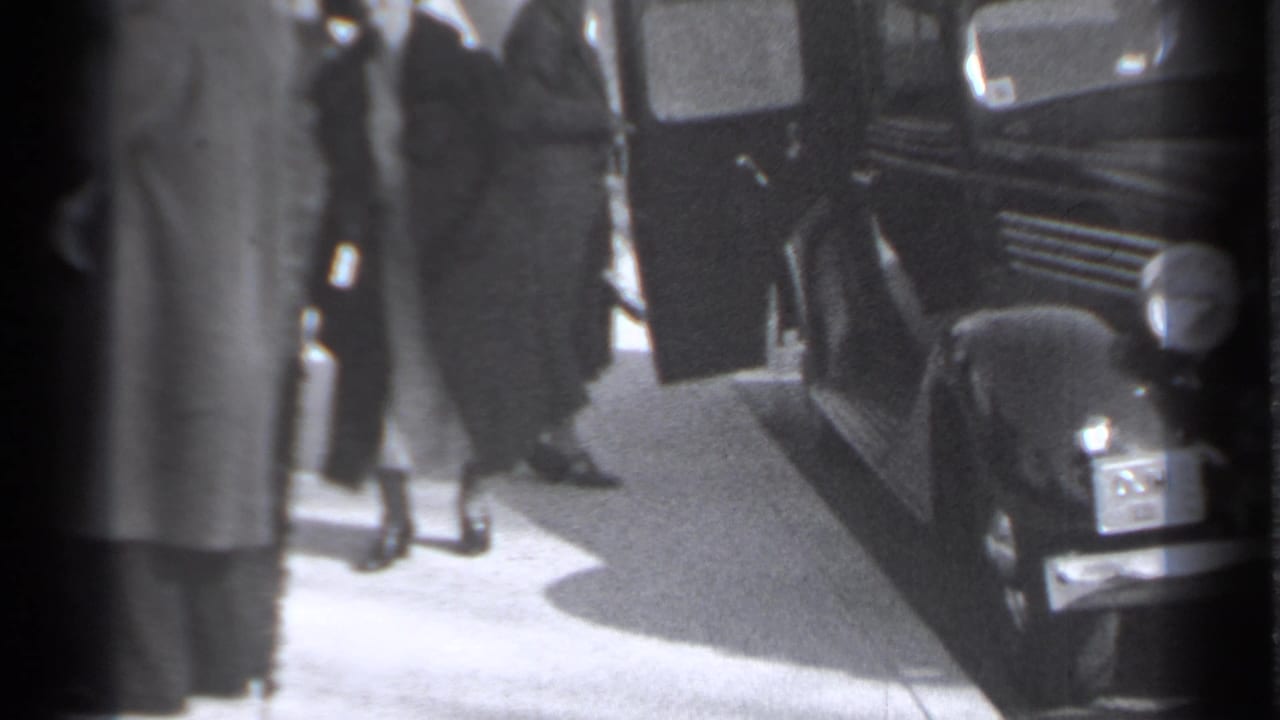 Woman in a long coat steps near a dark vintage automobile on a sidewalk.