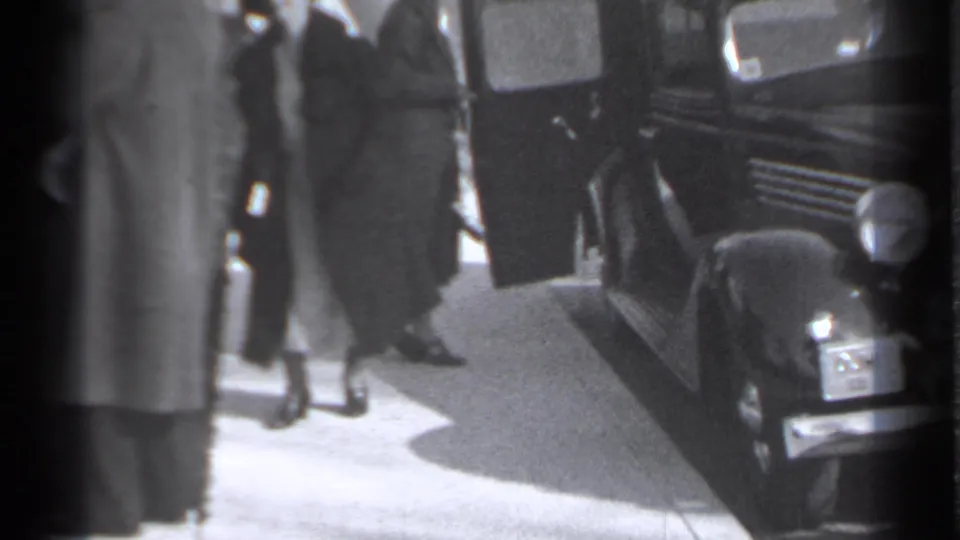 In 1939 New York home-movie footage, a smartly dressed woman stands beside a dark vintage automobile as other adults gather nearby.