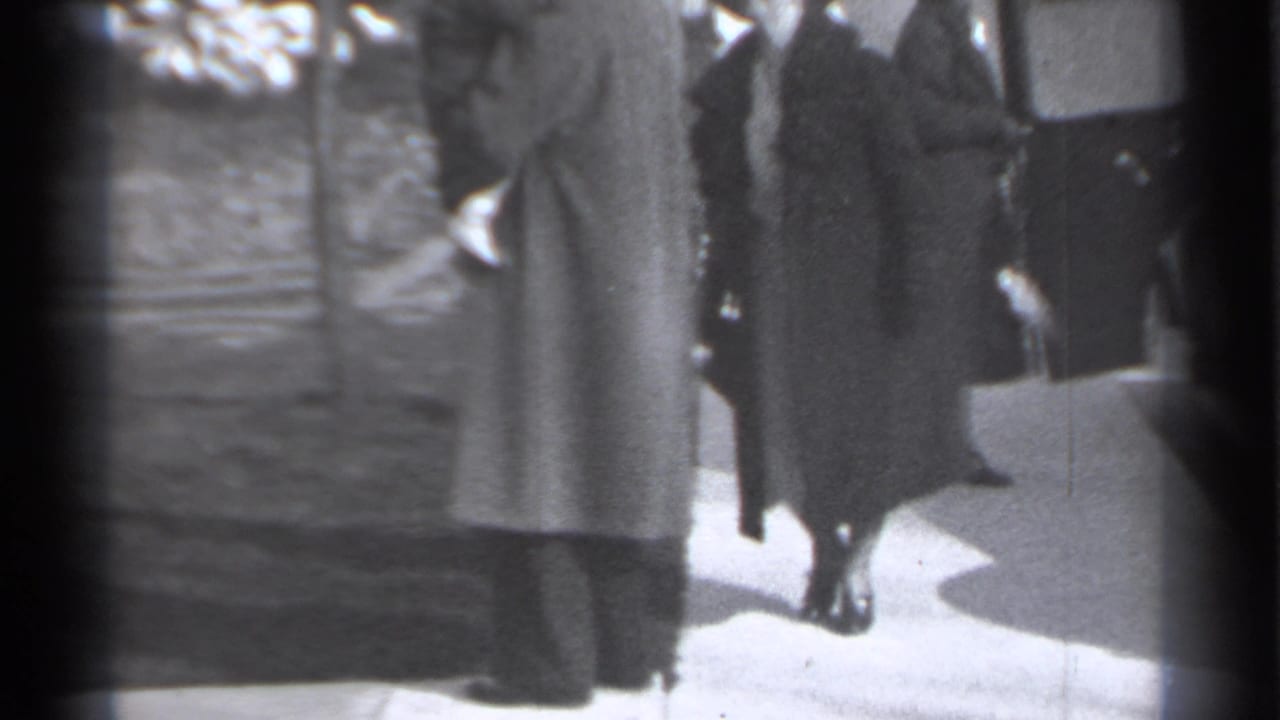 Adults in long coats stand near a sidewalk and bare ground beside a vehicle.