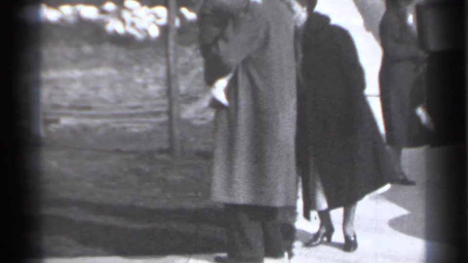 A woman in a dark coat walks toward a taller man in a long coat on a New York sidewalk, with another adult partly visible nearby.