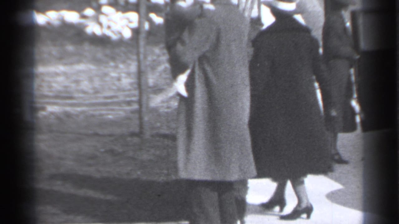 Two women in long coats stand on a sidewalk beside a planted area.