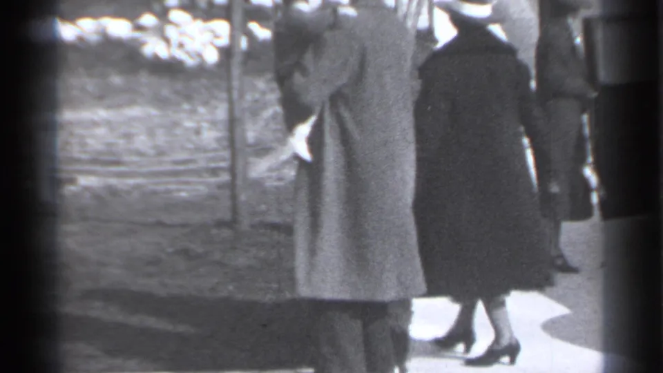 In 1939 New York home-movie footage, two women in long coats and hats stand near a sidewalk and planted strip.