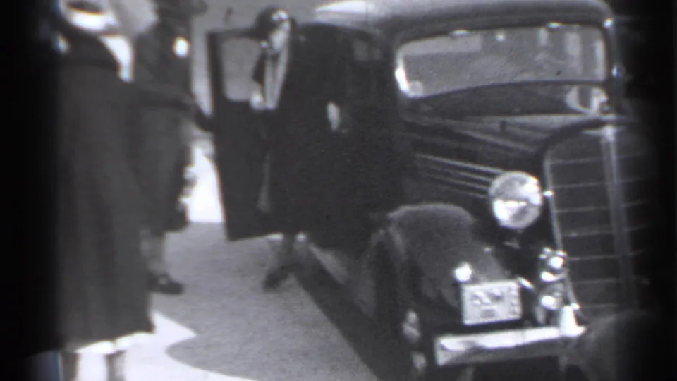 1939 New York home-movie frame showing a woman near the front of a dark classic automobile on a sidewalk.