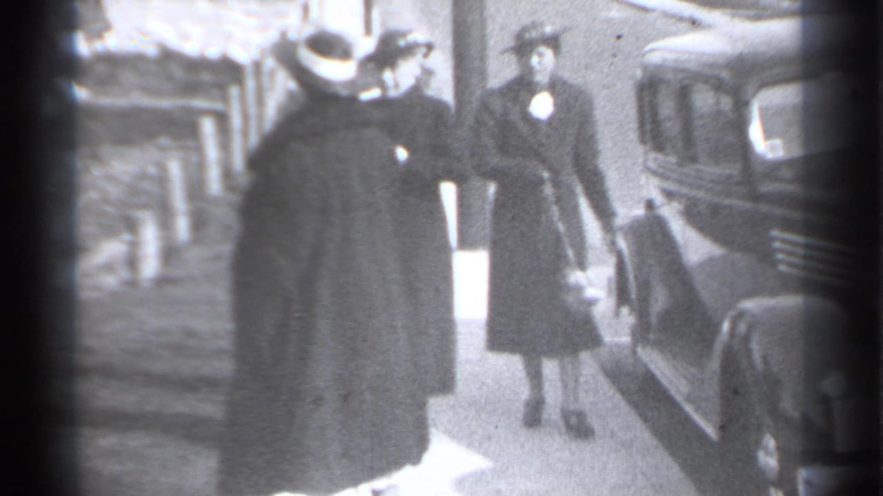 Two women and a suited man stand near a classic car on a sidewalk.