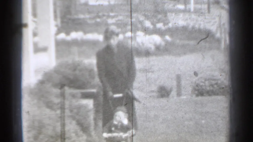 Black-and-white 8mm frame from New York in 1939 showing a suited man bending near a small object by a garden path.