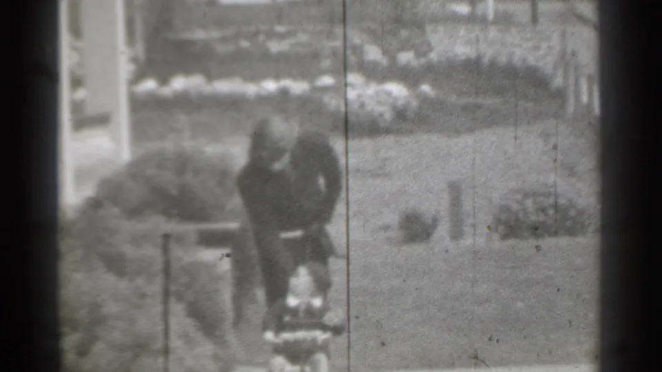 1939 New York home-movie frame showing a suited man bending in a yard area near a sidewalk and planted border.