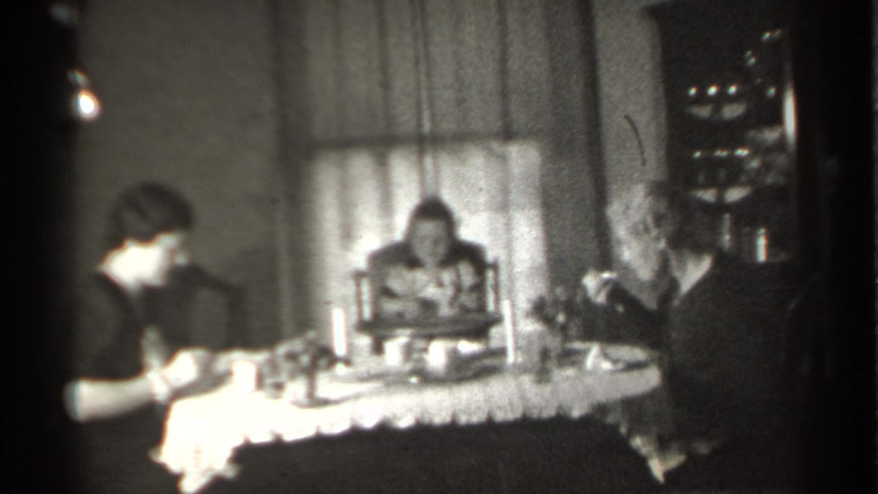 Three people seated at a dining table in a black-and-white home movie frame.