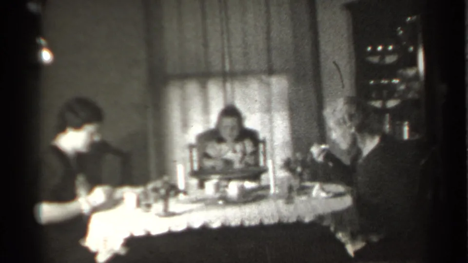 1939 New York home-movie frame showing two adults and a young child seated around a dining table in a curtained room.