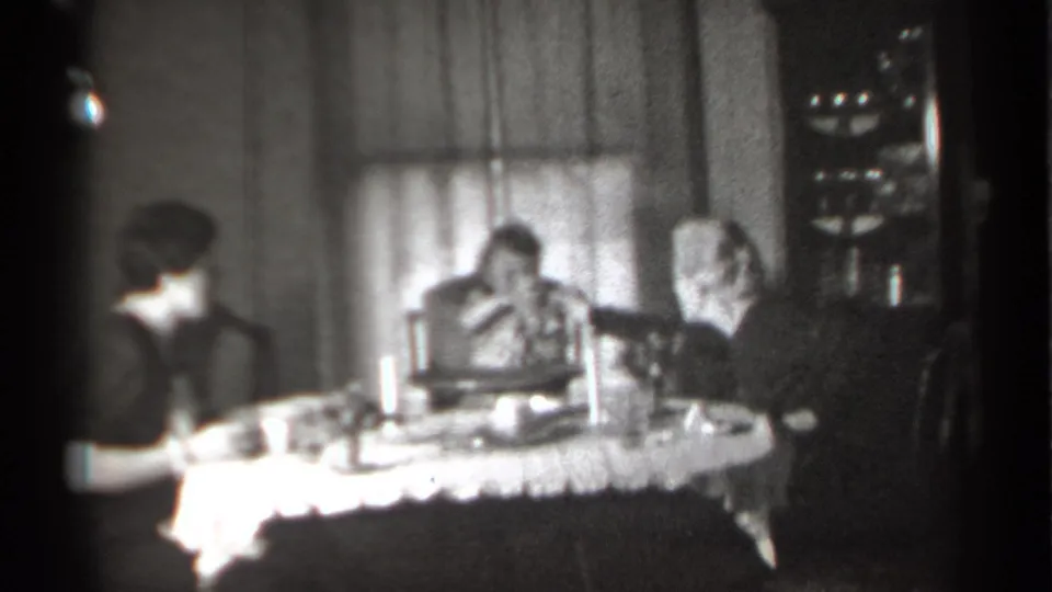 Black-and-white home-movie frame from 1939 New York showing an older woman reaching toward a child at the dinner table.