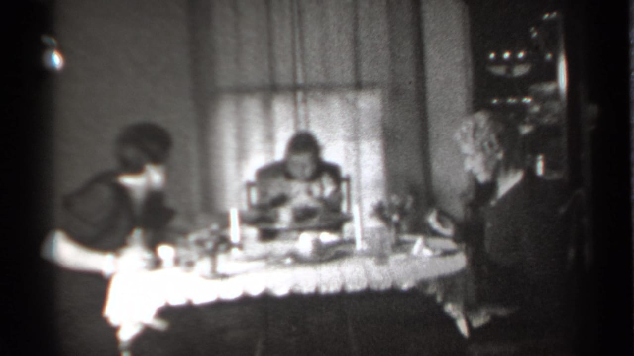 Two women and a small girl sit at a lace-covered dinner table in a dim home interior.
