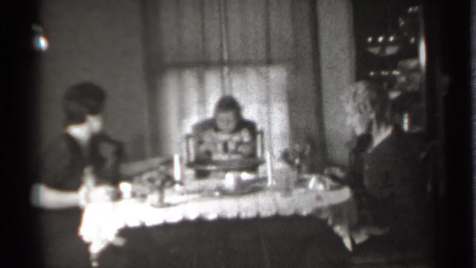 1939 New York domestic frame with a small girl seated between two women at a lace-covered table in soft black-and-white 8mm footage.