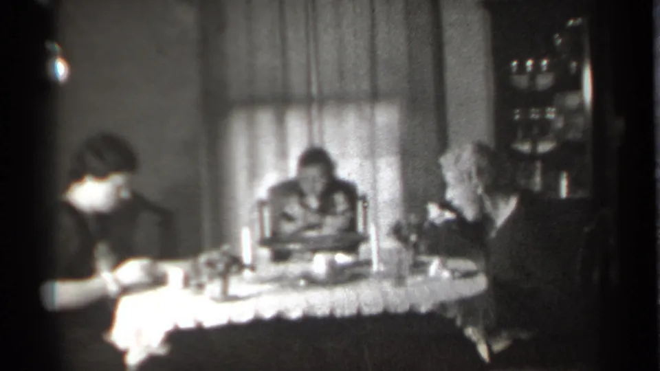 1939 New York home interior frame showing the dinner group from a slightly shifted angle, with tableware, a candle, and curtains behind them.