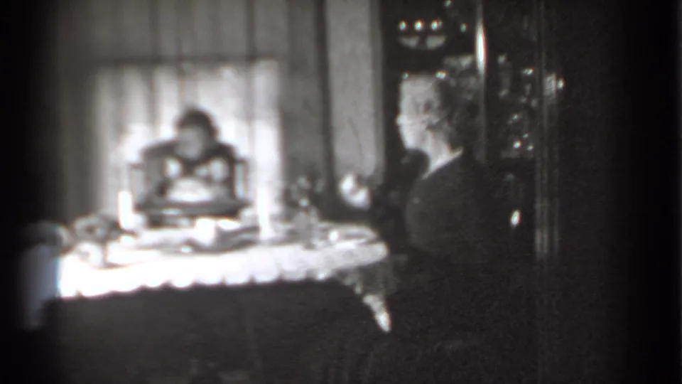 Soft black-and-white frame of a woman at a household table as a baby sits in a high chair near curtained windows.