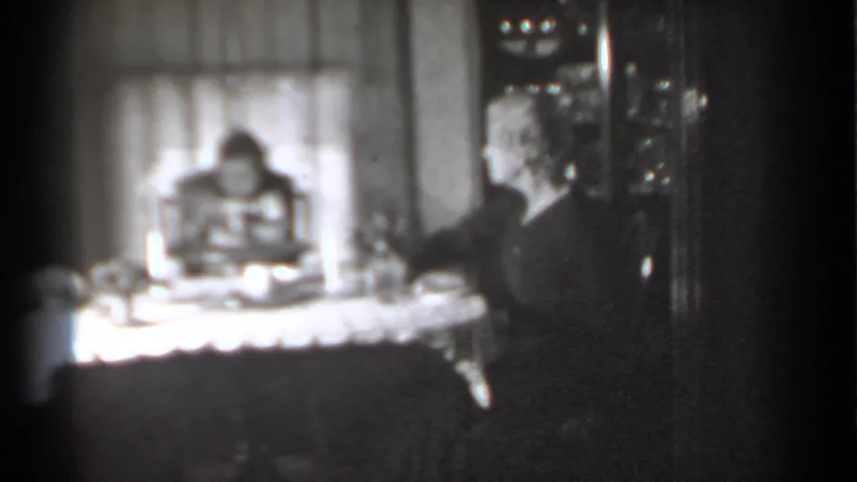 Grainy 8mm frame showing the woman extending an arm near the table while the baby remains seated in the high chair.