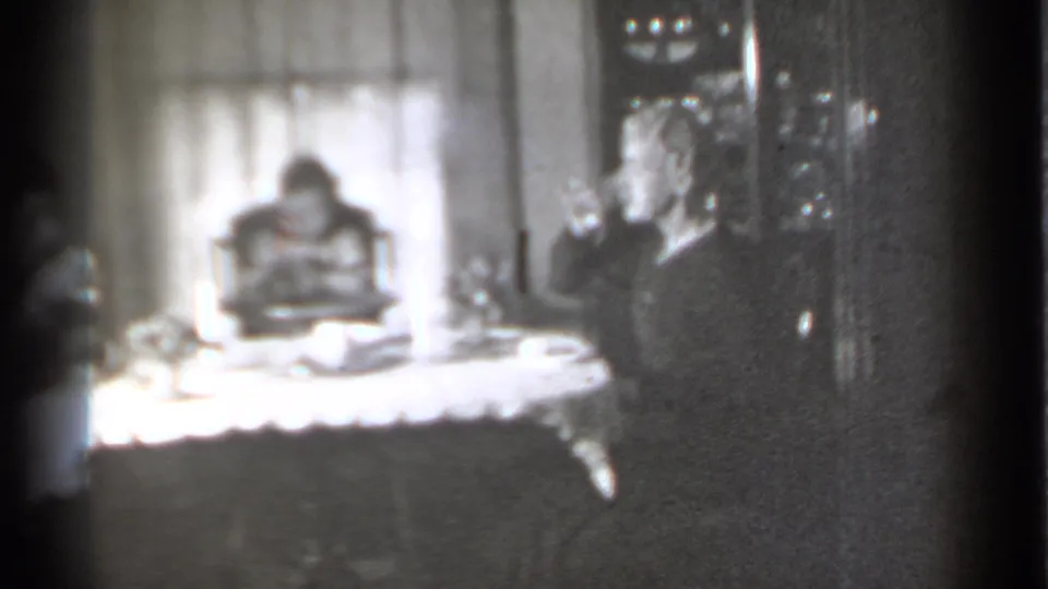 Black-and-white home-movie frame with the woman lifting a hand near her mouth and the baby seated in a high chair beyond the table.