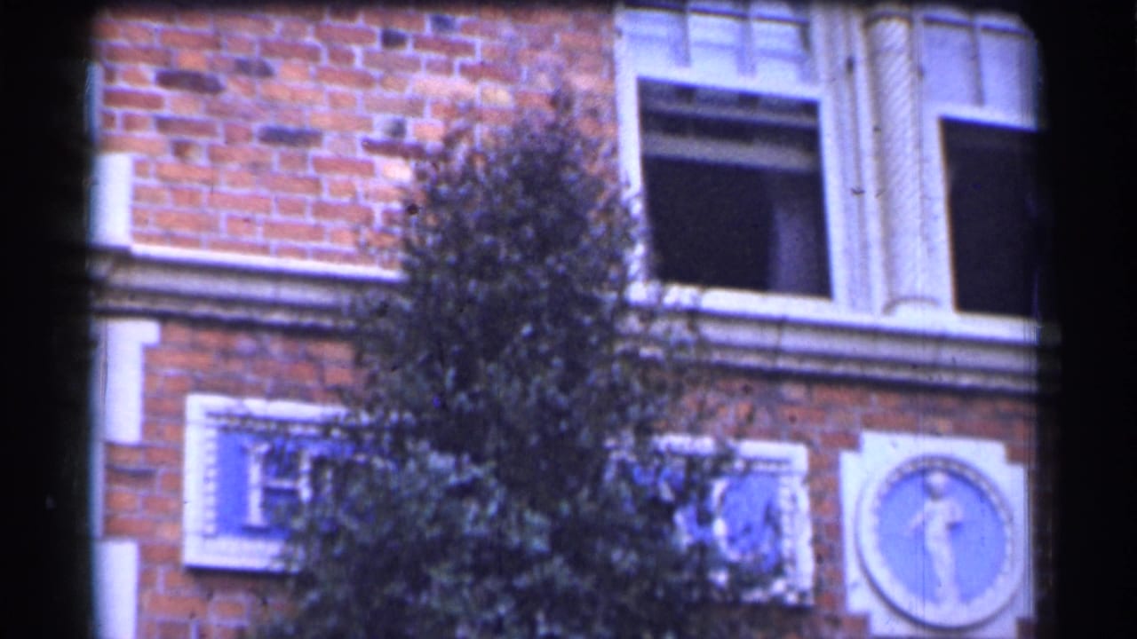 1944 San Francisco hospital exterior with red brick, a leafy tree, blue plaques, and a circular figure emblem partly obscured by branches.