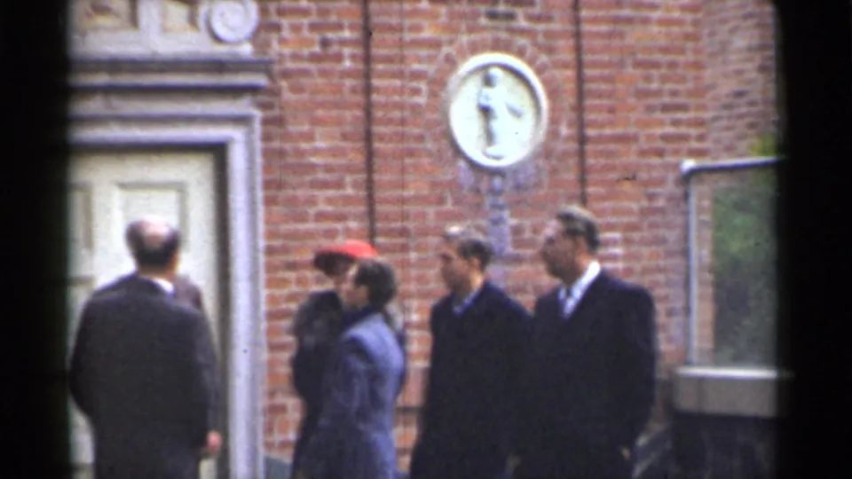 Adults in mid-century coats and suits stand outside a San Francisco brick building, with a wall plaque visible above.