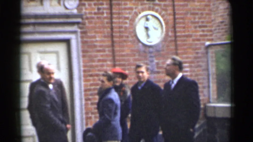 1944 San Francisco exterior frame with adults in suits and coats conversing beside a brick building entrance.