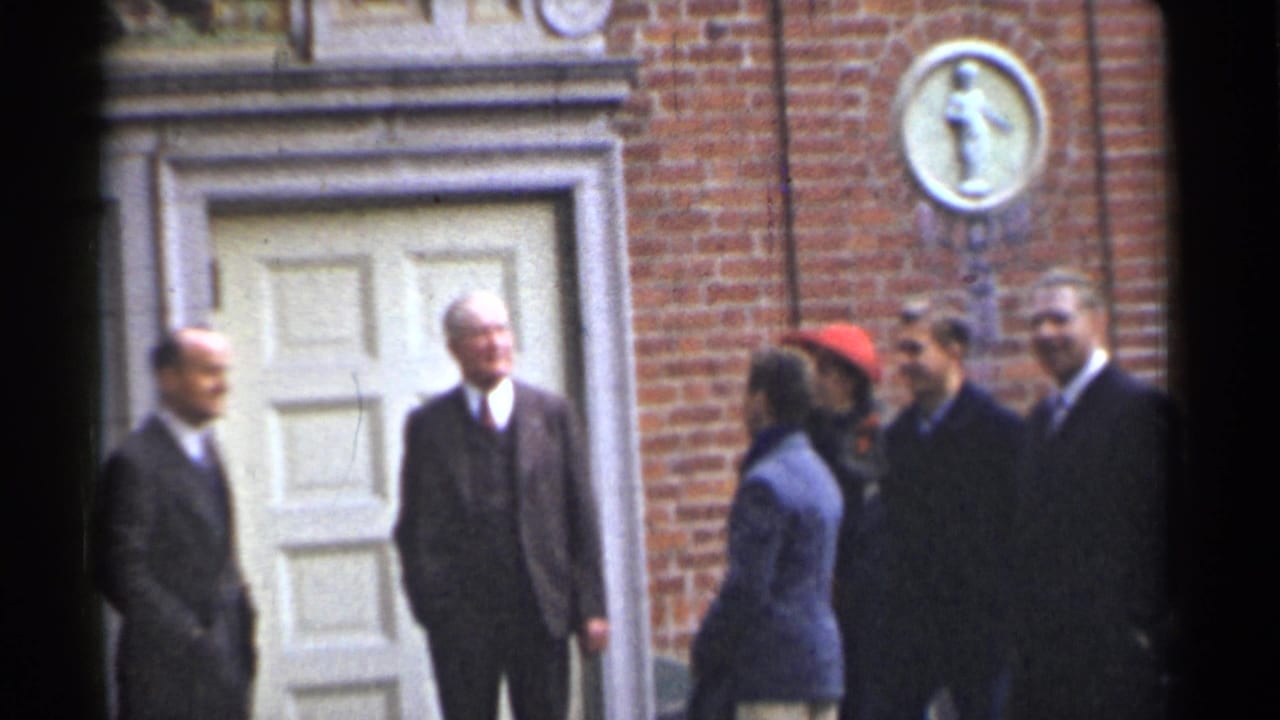 Adults in suits and coats stand outside a brick building with a white door.