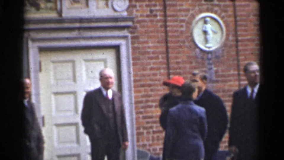 Adults in period clothing stand close together outside the brick facade, including a woman in a red hat and men in dark suits.