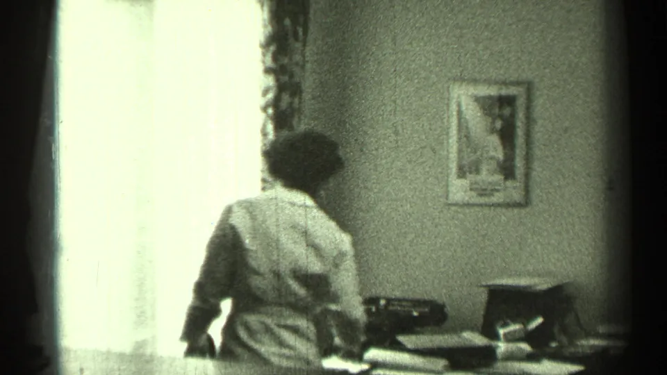 A brief interior view shows a person by a bright window with a desk, typewriter-like object, papers, and framed wall art.
