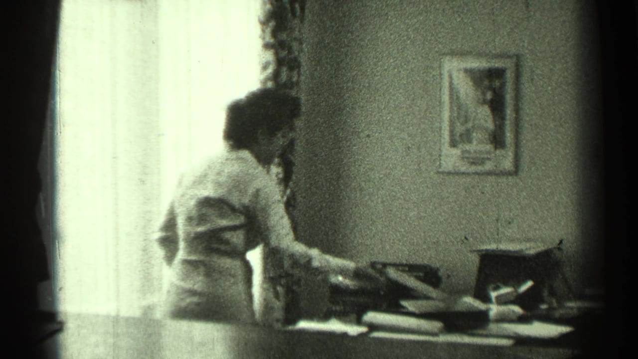 Woman standing or leaning beside a desk with a typewriter near a bright curtained window.