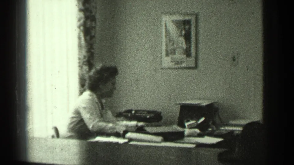 The woman sits at a desk in the San Francisco interior, facing a typewriter with papers spread across the work surface.
