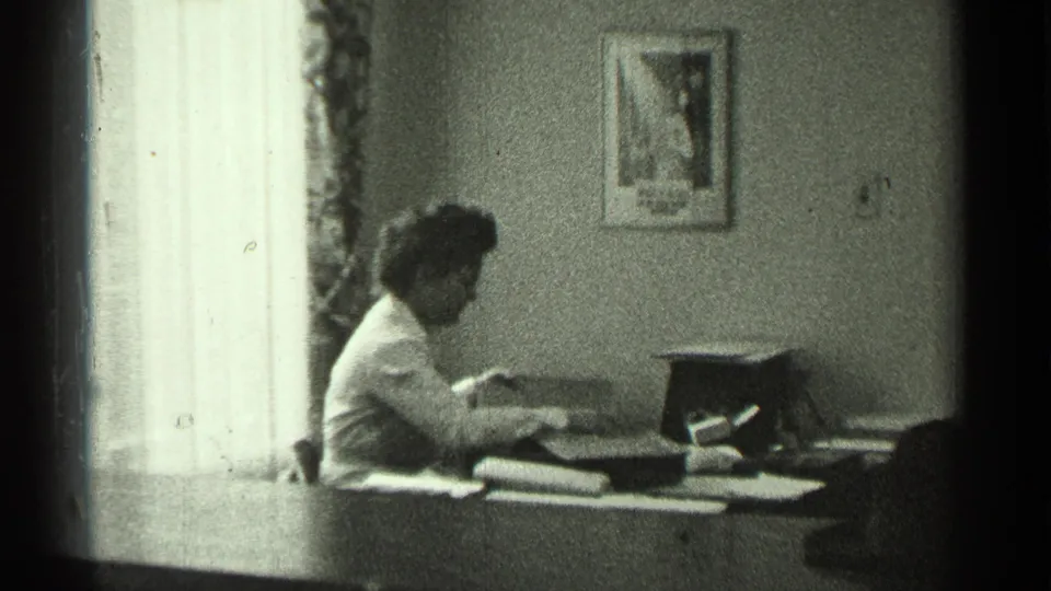 The woman appears to read or handle papers at the desk, with a typewriter, stacked materials, and the bright window still visible.