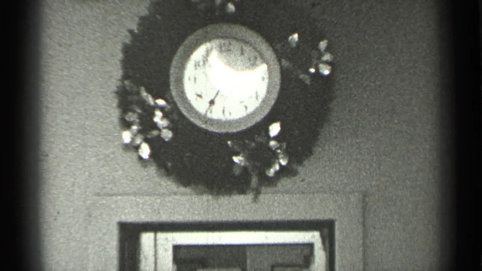 The camera holds on the wreath-framed analog clock, with doorway trim and soft 8mm grain visible below.