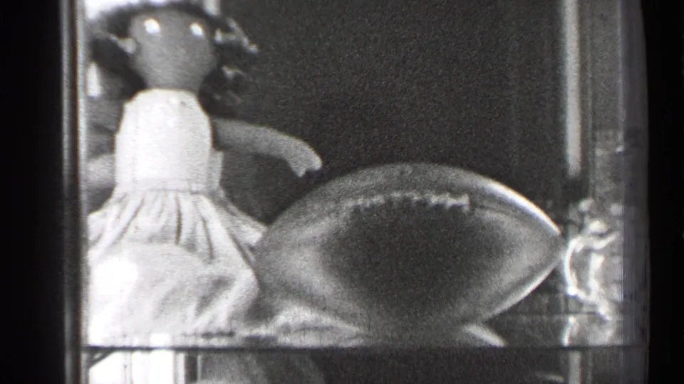 1944 San Francisco 8mm frame showing a glass case with a dressed doll, football, shelves, and reflected highlights.