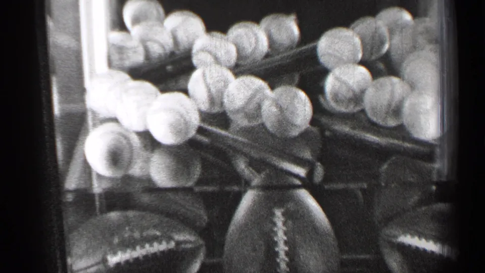 Black-and-white 8mm view of baseballs, footballs and trophy forms in a San Francisco sports display case.