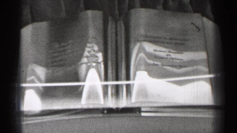 Black-and-white 8mm view of a glass-covered book display in San Francisco, with printed text and a small artifact-like object visible.