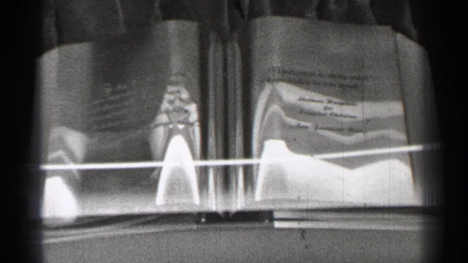1944 black-and-white home-movie frame of an open book behind glass, showing page text, a small central object, and dense film texture.