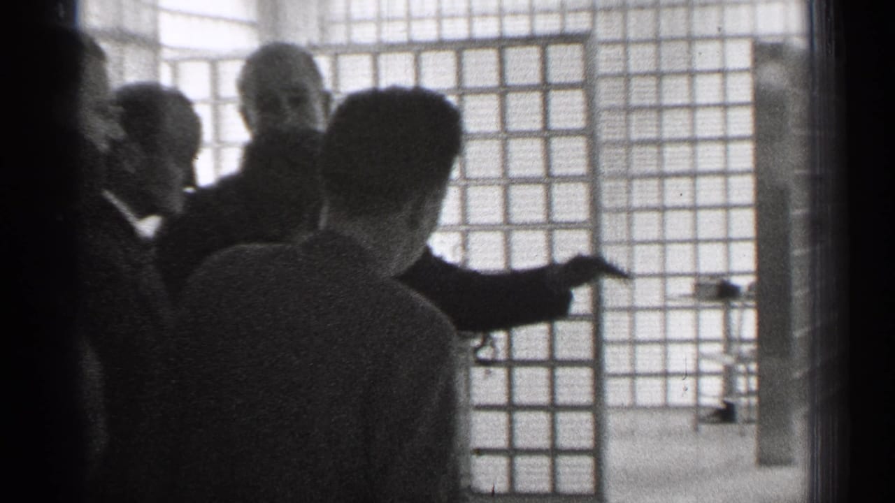 In 1944 San Francisco home-movie footage, adults in formal clothing watch a suited man gesture beside a grid-patterned interior window or partition.