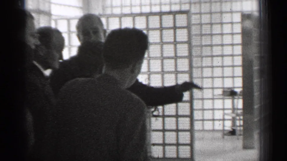 In 1944 San Francisco home-movie footage, adults in formal clothing watch a suited man gesture beside a grid-patterned interior window or partition.