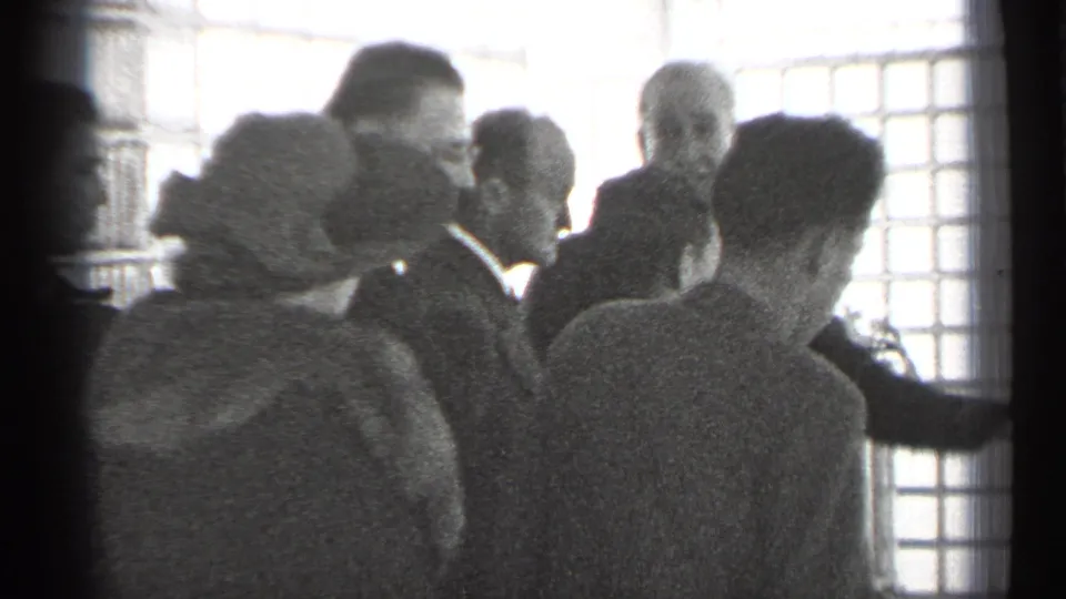 Adults in 1940s formal attire stand closely together near bright grid-patterned windows during an indoor San Francisco presentation or tour.