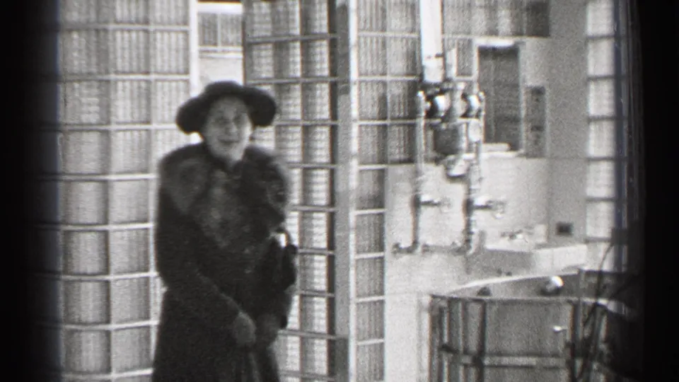 In 1944 San Francisco home-movie footage, a woman in a hat and fur-collared coat stands near glass block walls in a sunlit interior.