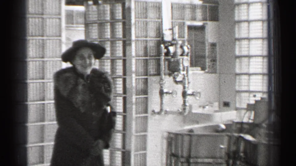 A woman in 1940s San Francisco footage smiles toward the camera while standing in a bright interior with glass block walls.