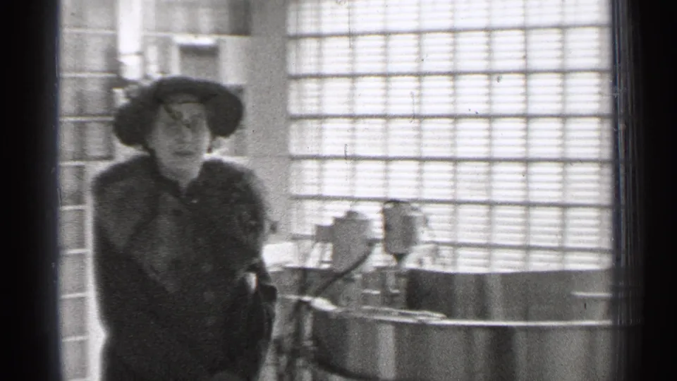 The woman stands closer to the camera in the San Francisco home-movie scene, with glass blocks and a bright interior behind her.