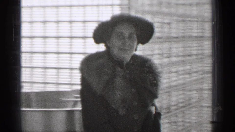 A soft 8mm close view shows the woman in a hat and fur-collared coat against a bright windowed or glass block wall.